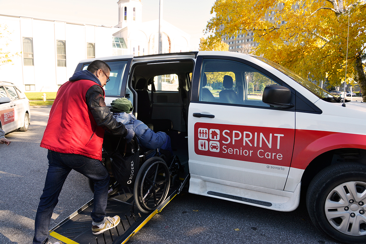Senior na wózku inwalidzkim zostaje popchnięty do furgonetki Sprint Senior Care przez człowieka w czerwonej kamizelce